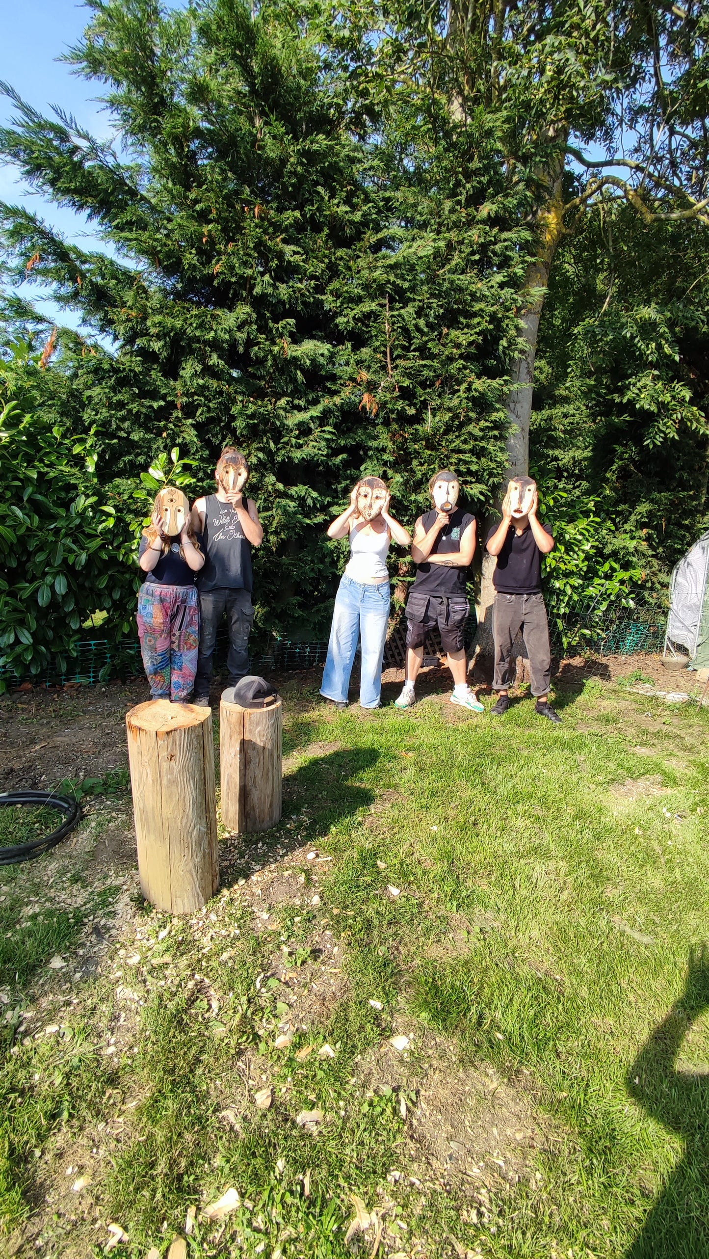 Five people outdoors proudly wearing the masks the have made in a garden with trees and grass.