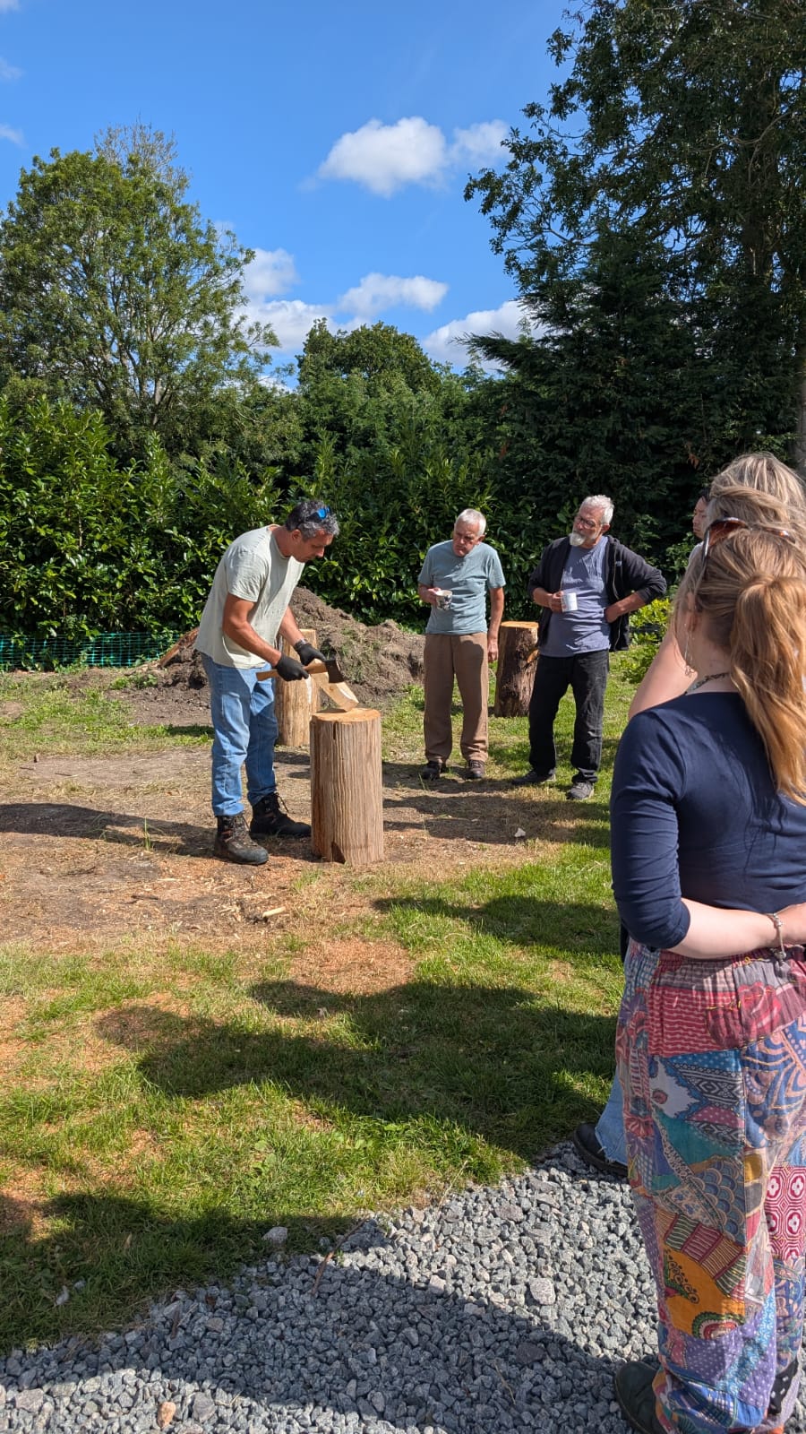 People gathered around, listening to instructions on how to carve a mask using an axe on a wooden block outdoors.